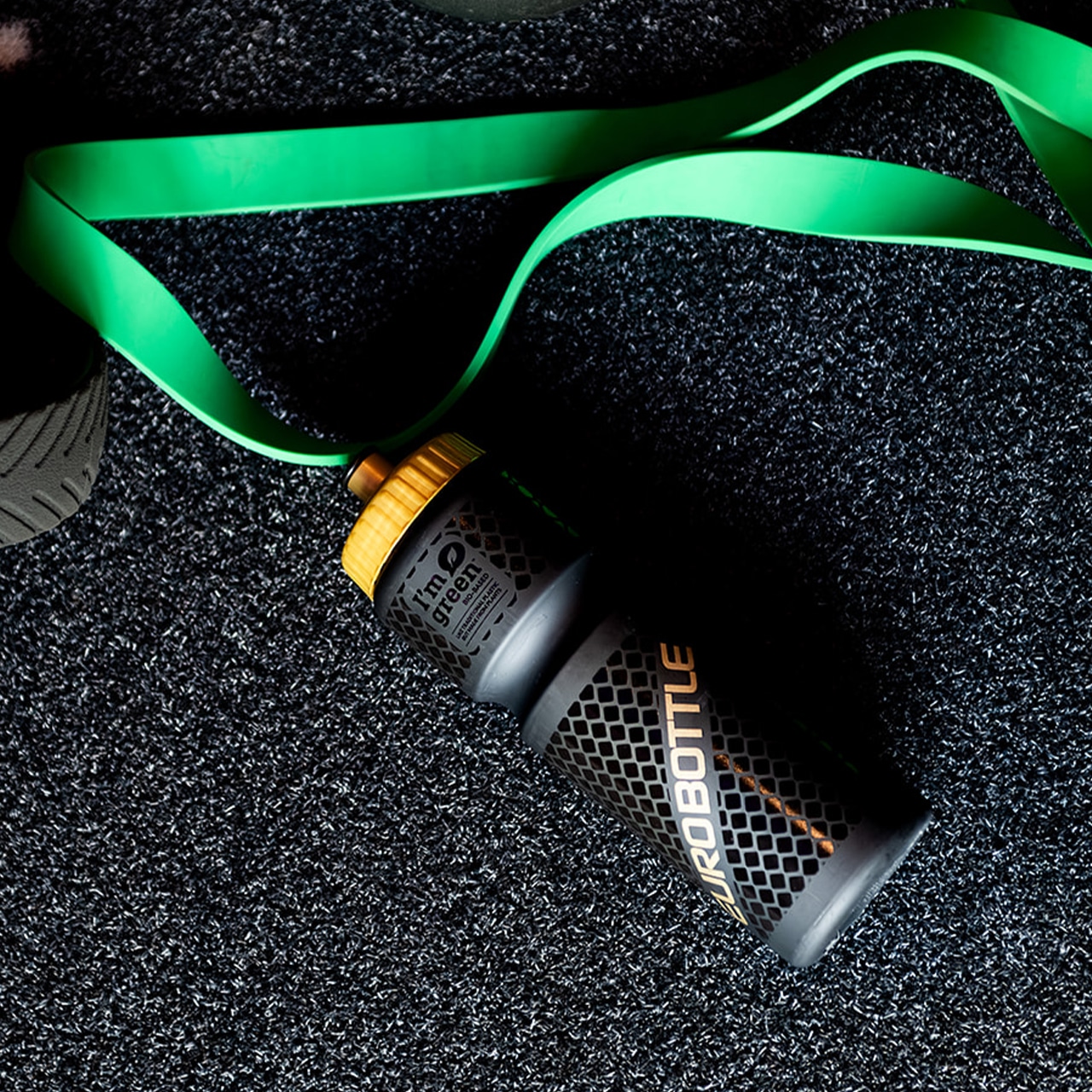 Zwarte Eurobottle bidon met gele dop op sportvloer naast groene weerstandsband