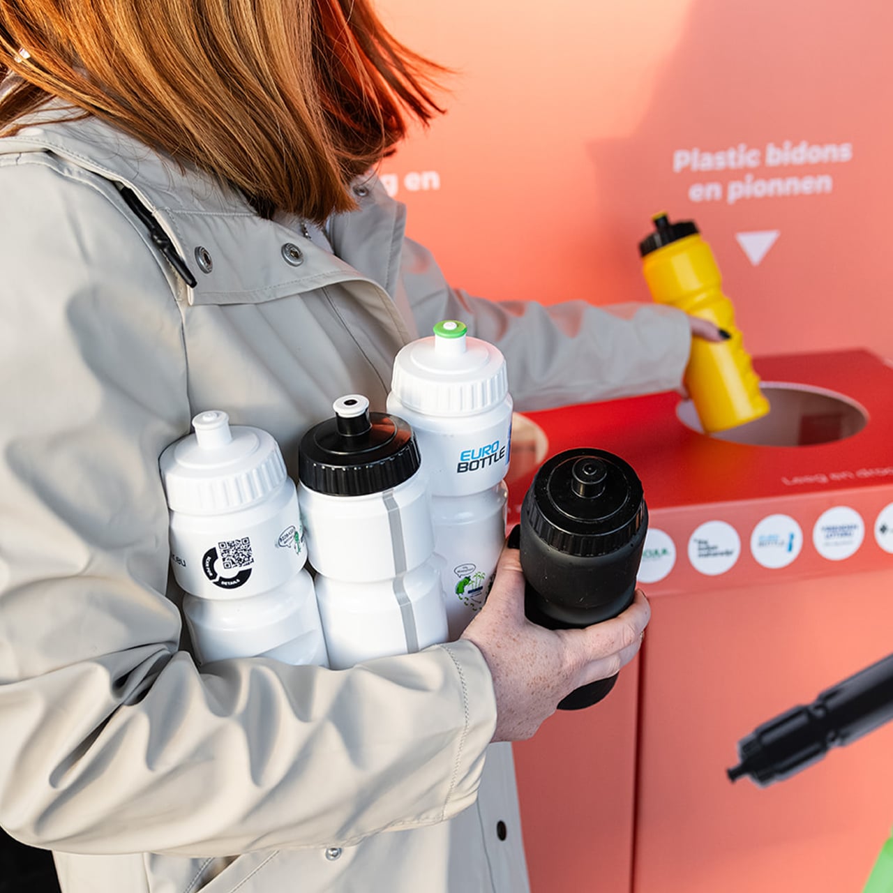 Een vrouw levert verschillende gebruikte Eurobottle bidons in bij een ClubHub-bak, waar QR-codes op de flessen verwijzen naar het productpaspoort en de juiste verwerking
