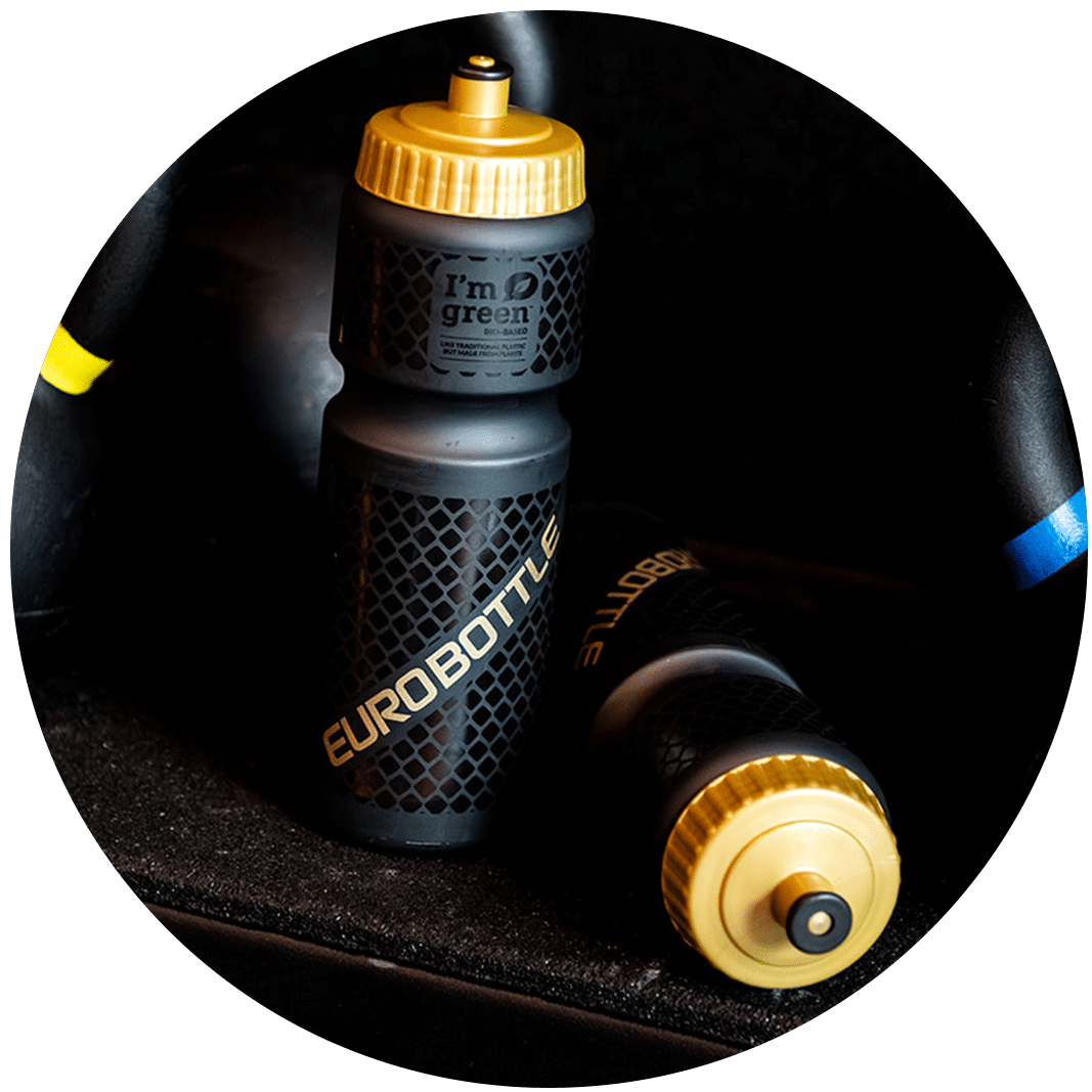 Zwarte Signature bidon van Eurobottle met goudkleurige dop en reliëfstructuur, gefotografeerd in een sportomgeving tussen kettlebells.