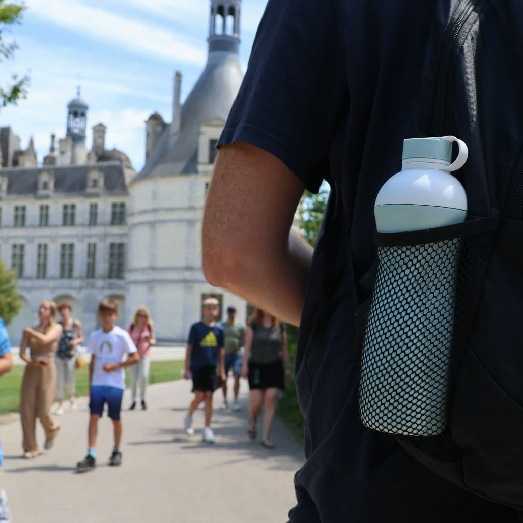 Weiße Oasus-Trinkflasche in der Seitentasche eines Rucksacks bei einem wandernden Touristen vor einem Schloss, nachhaltige Flasche für unterwegs