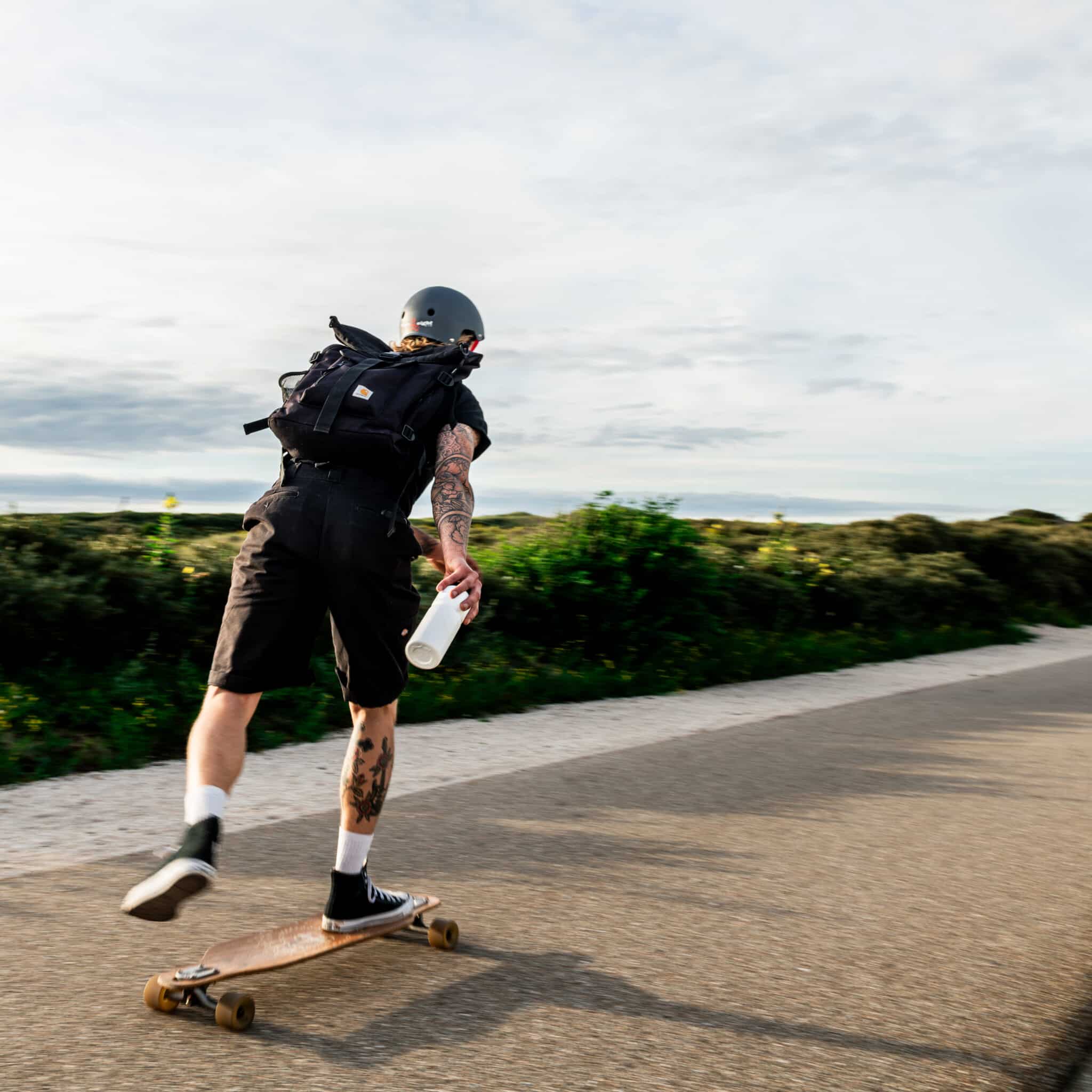 Ein Longboarder mit einer Trinkflasche auf einem Weg am Wasser entlang.