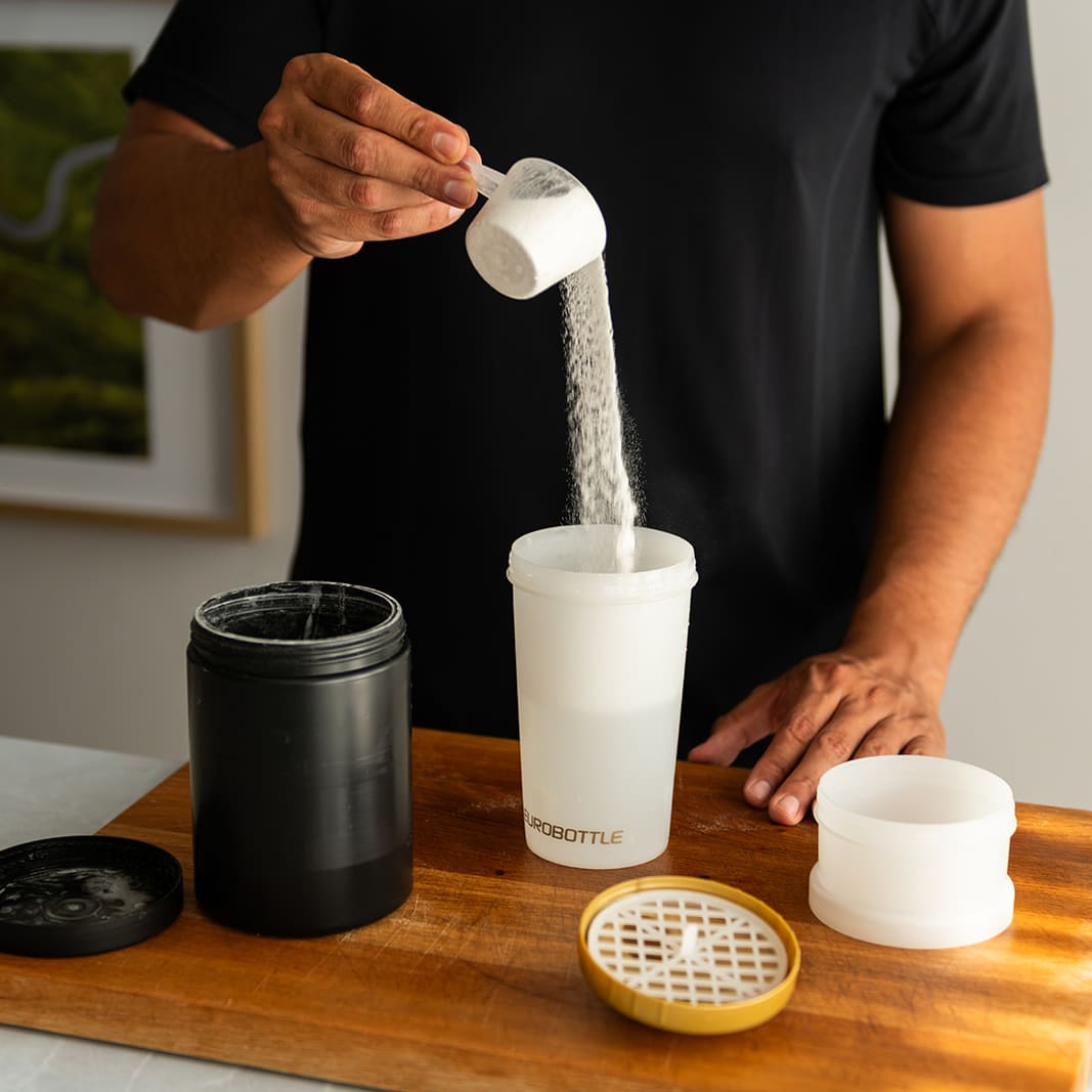 Man voegt eiwitpoeder toe aan een witte Eurobottle shaker van 550 ml, met het mengrooster ernaast